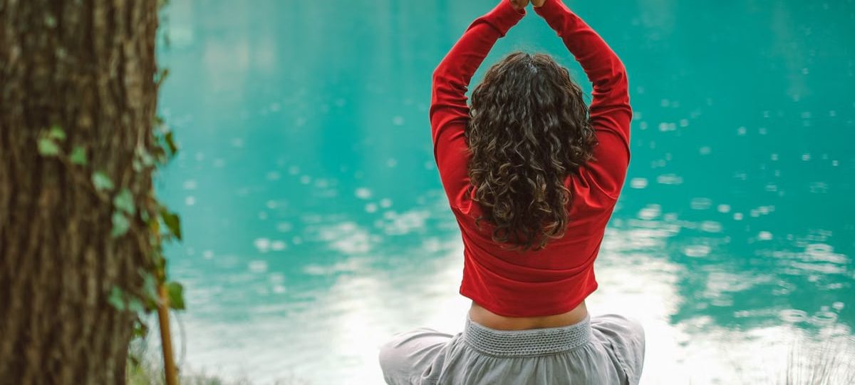 A woman meditates in nature on a rock by a tranquil turquoise lake, practicing yoga.