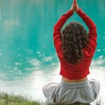 A woman meditates in nature on a rock by a tranquil turquoise lake, practicing yoga.