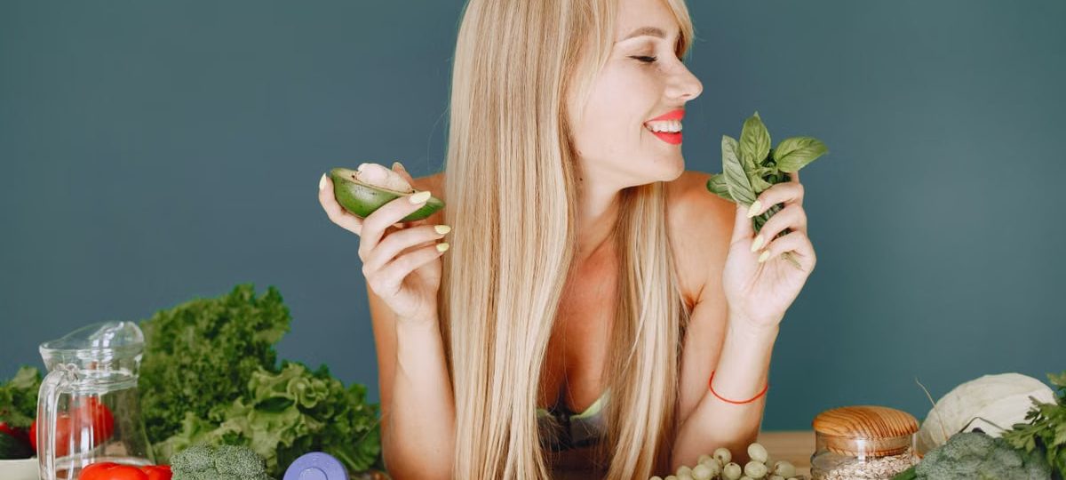 A smiling woman holding avocado and basil, surrounded by fresh vegetables and fruits for healthy eating.
